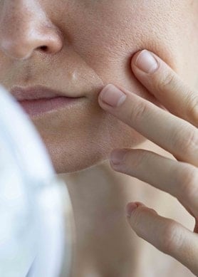 Frau berührt die Wange und prüft ihre großporige Haut im Spiegel