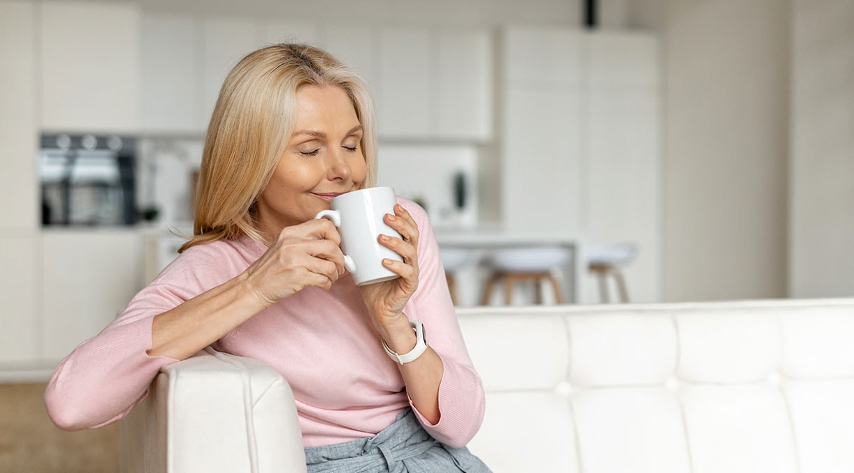 Frau mittleren Alters sitzt mit geschlossenen Augen und einer Tasse Tee auf einer Couch 