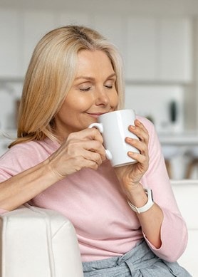 Frau mittleren Alters sitzt mit geschlossenen Augen und einer Tasse Tee auf einer Couch