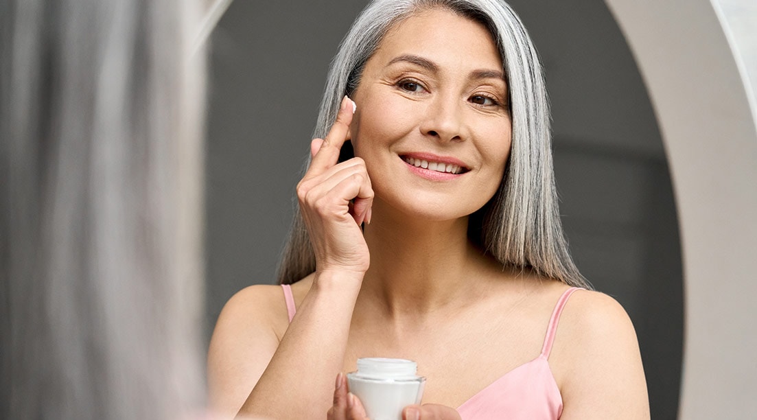 Frau mittleren Alters steht vor dem Spiegel und trägt Creme auf