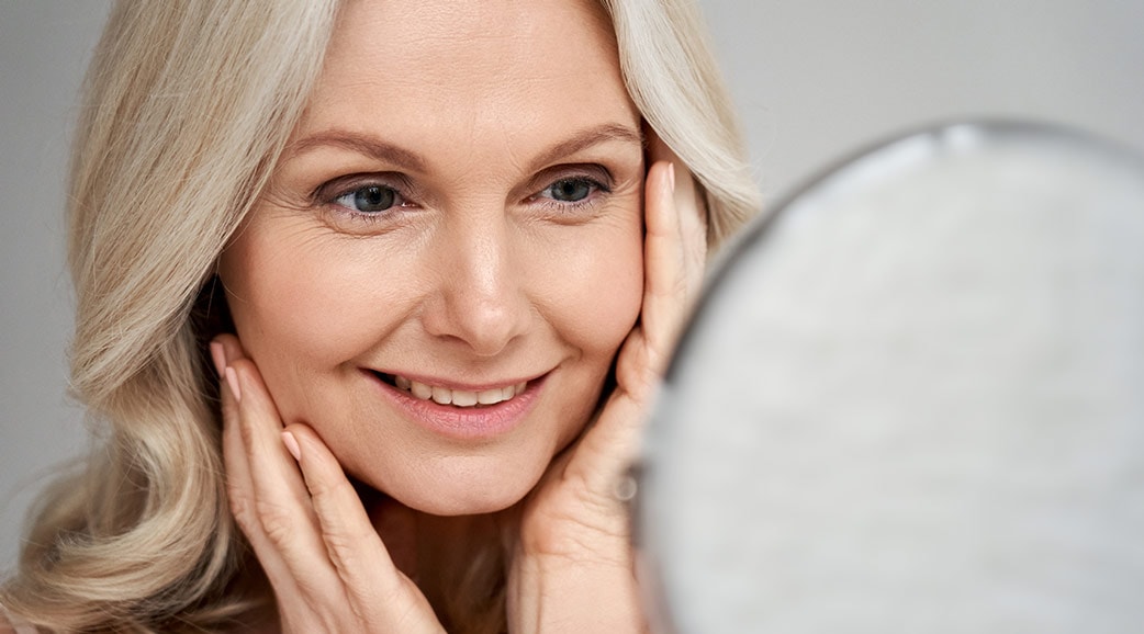 Eine lächelnde Frau mittleren Alter mit ihren Händen an den Wangen betrachtet sich im Spiegel 