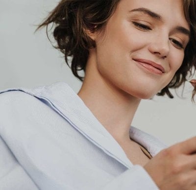 Person in weißem Hemd hält ein Smartphone in der Hand und berührt mit der anderen Hand eine Haarsträhne.