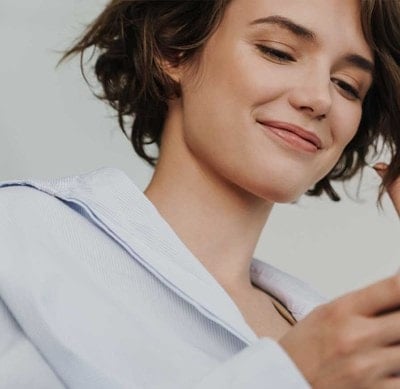 Person in weißem Hemd hält ein Smartphone in der Hand und berührt mit der anderen Hand eine Haarsträhne.
