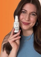 Person hält eine Vichy Capital Soleil Sonnenpflege-Flasche vor orangefarbenem Hintergrund, daneben zwei weitere Produkte aus der gleichen Serie.