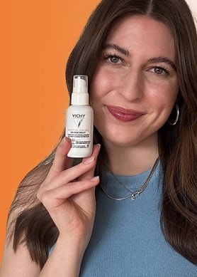 Person hält eine Vichy Capital Soleil Sonnenpflege-Flasche vor orangefarbenem Hintergrund, daneben zwei weitere Produkte aus der gleichen Serie.