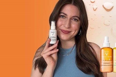 Person hält eine Vichy Capital Soleil Sonnenpflege-Flasche vor orangefarbenem Hintergrund, daneben zwei weitere Produkte aus der gleichen Serie.