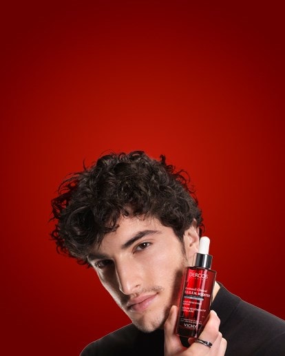 Mann mit lockigem Haar hält eine rote Flasche Vichy Dercos Regen Anti-Haarausfall-Serum für empfindliche Kopfhaut vor rotem Hintergrund.