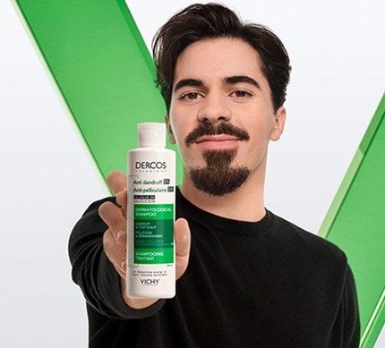 Person hält eine Flasche Vichy Dercos Anti-Schuppen-Shampoo für empfindliche Kopfhaut vor grünem Hintergrund in die Kamera