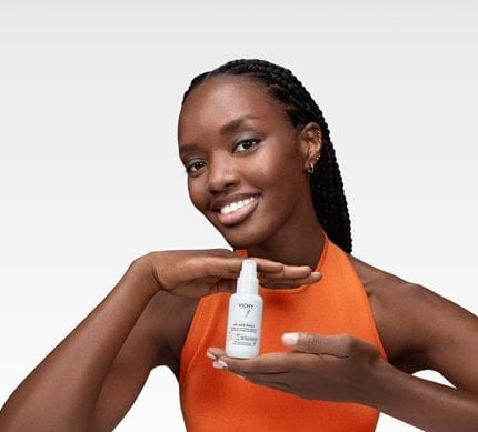 Frau in orangefarbenem Oberteil hält lächelnd ein Vichy UV-Age Daily Sonnenschutz mit Lichtschutzfaktor 50 in beiden Händen vor einem weißen Hintergrund