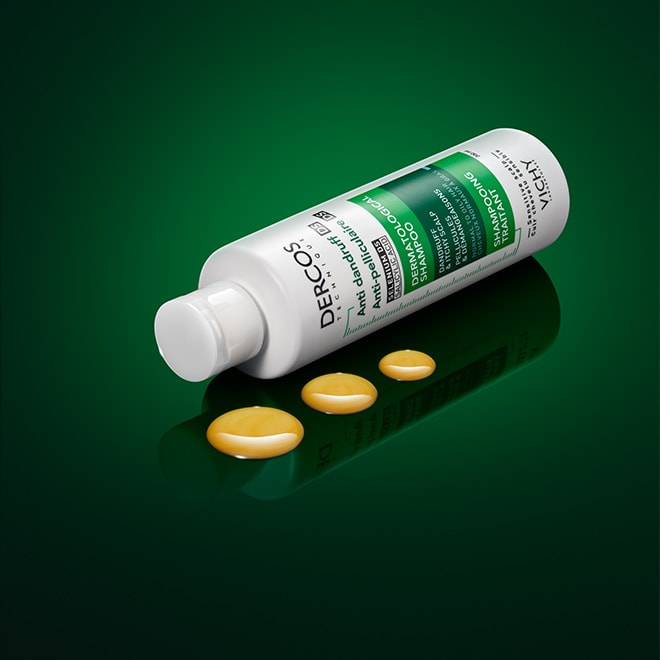 Flasche des Vichy Dercos Anti-Schuppen Shampoos für normales bis fettiges Haar, liegend auf grünem Hintergrund, mit gelblicher Shampoo-Textur in drei Tropfen daneben