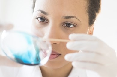 Eine Person in einem Labor hält eine Petrischale mit blauer Flüssigkeit, die von lichtdurchfluteter Umgebung umgeben ist.