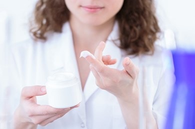 Frau testet weiße Creme aus einem Tiegel auf ihrem Finger, Hintergrund mit Laborblau und unscharfen Details.