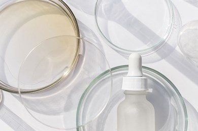 Glasfläschchen mit Pipette, umgeben von transparenten Petrischalen und Glasgefäßen auf weißem Hintergrund, hell ausgeleuchtet.