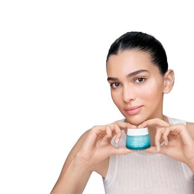 Person hält einen Cremetiegel von Vichy Mineral 89 in den Händen, trägt ein weißes Oberteil und präsentiert das Produkt vor hellem Hintergrund.