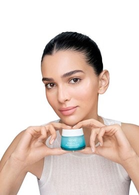 Person hält einen Cremetiegel von Vichy Mineral 89 in den Händen, trägt ein weißes Oberteil und präsentiert das Produkt vor hellem Hintergrund.
